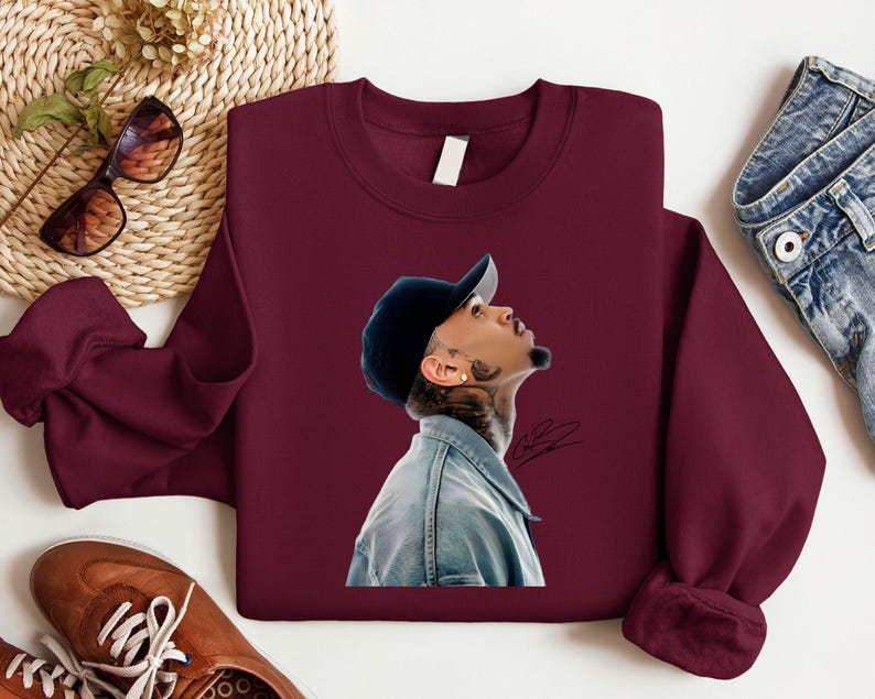 May include: Burgundy crewneck sweatshirt featuring a portrait of a person wearing a denim jacket and a black baseball cap. The portrait is on the front of the sweatshirt. Sunglasses and brown leather shoes are visible in the background.