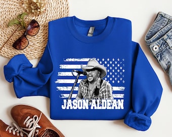 May include: Royal blue sweatshirt featuring a graphic of a man wearing a cowboy hat in front of an American flag design. The text "JASON ALDEAN" is printed in white below the graphic. The sweatshirt is made of a soft material.