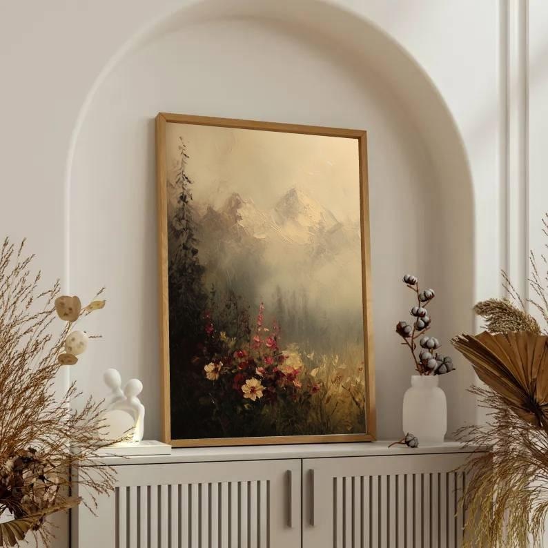 May include: A framed landscape painting with a gold-colored frame. The artwork depicts a mountain scene with a forest and colorful flowers in the foreground. The painting is displayed on a white cabinet.