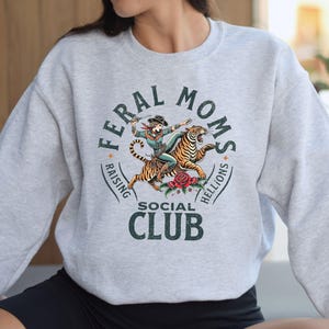 May include: A gray sweatshirt with a graphic print of a woman riding a tiger. The graphic includes the text "Feral Moms Raising Hellions Social Club".