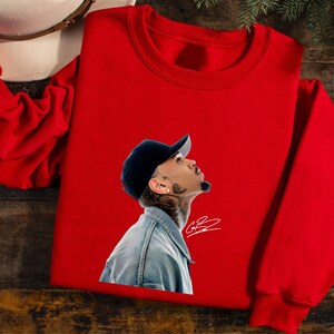 May include: A red crewneck sweatshirt featuring a graphic of a person wearing a denim jacket and a black cap. The graphic includes a signature. A white hat is visible in the upper left corner.
