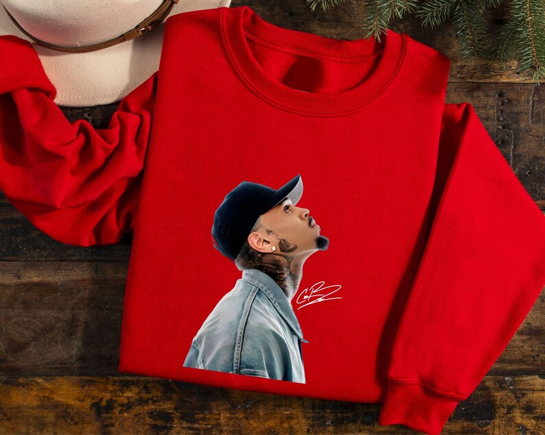 May include: A red crewneck sweatshirt featuring a graphic of a person wearing a denim jacket and a black cap. The graphic includes a signature. A white hat is visible in the upper left corner.