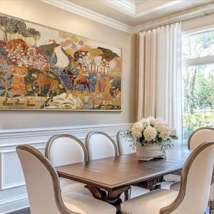 May include: An ornate framed painting depicting a historical scene hangs above a wooden dining table. The table is surrounded by white upholstered chairs. A vase of white flowers sits in the center of the table, and white curtains frame a window.