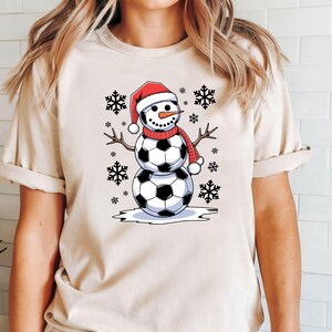 May include: A beige t-shirt with a graphic of a snowman made of soccer balls wearing a red Santa hat and a red scarf. The snowman is surrounded by snowflakes.