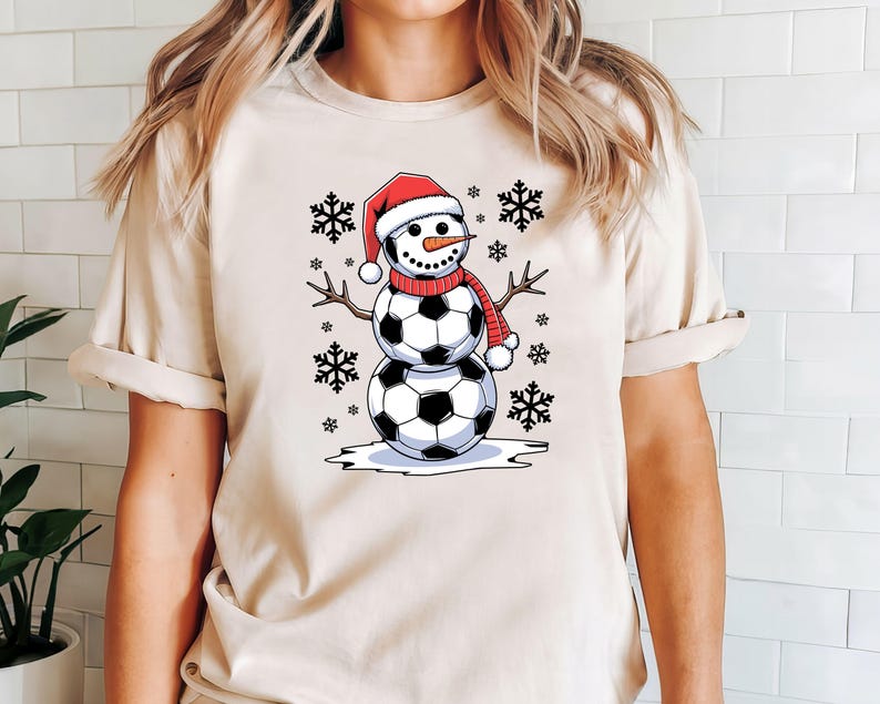 May include: A beige t-shirt with a graphic of a snowman made of soccer balls wearing a red Santa hat and a red scarf. The snowman is surrounded by snowflakes.