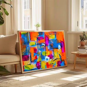May include: Abstract art print with a wooden frame. The artwork features a vibrant mix of colors, including blue, orange, yellow, and pink, with black lines. The painting is displayed on a rug in a well-lit room.