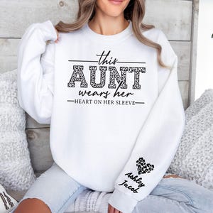 May include: White sweatshirt with black text that reads "This Aunt wears her heart on her sleeve". The text is surrounded by a leopard print design. The sleeve has a black heart with the names "Ashley Jacob" written below it.