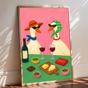 May include: A framed art print featuring two geese wearing sunglasses and accessories, enjoying wine and snacks. The artwork includes a bottle of wine, glasses of red wine, cheese, olives, and a box that says "SASSY". The background is pink and green.