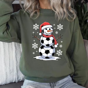 May include: A green sweatshirt with a snowman made of soccer balls wearing a red Santa hat. The snowman is surrounded by snowflakes.