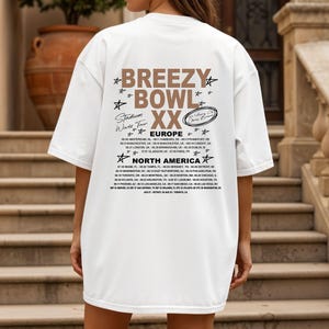 May include: White oversized t-shirt with the text "Breezy Bowl XX Stadium World Tour" in brown and black. The back of the shirt lists tour dates for Europe and North America. The shirt is worn by a person standing on a set of stairs.