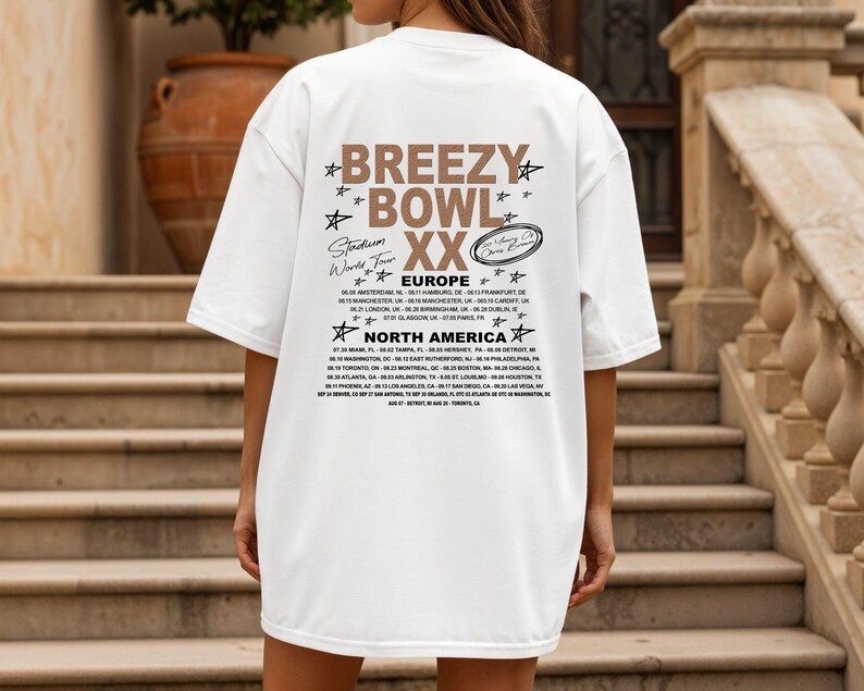May include: White oversized t-shirt with the text "Breezy Bowl XX Stadium World Tour" in brown and black. The back of the shirt lists tour dates for Europe and North America. The shirt is worn by a person standing on a set of stairs.