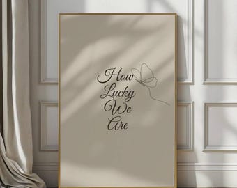 May include: A framed art print with a beige background and gold frame. The print features the phrase "How Lucky We Are" in elegant black script, accompanied by a delicate line drawing of a butterfly.