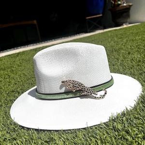 May include: White wide-brimmed straw hat with a green band and a gold leopard brooch.