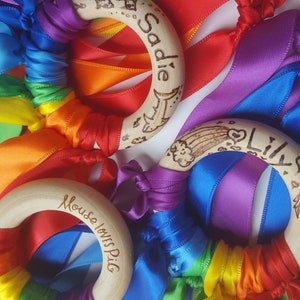 Op de afbeelding: Drie houten ringen met kleurrijke linten eraan bevestigd. De ringen zijn versierd met gepersonaliseerde tekst, waaronder "Sadie", "Lily" en "Mouse loves Pig".