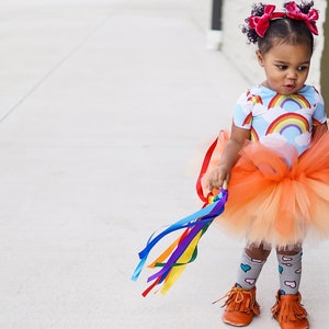 Op de afbeelding: Een jong meisje draagt een lichtblauw en wit regenboogprint onesie met een perzikkleurige tule tutu rok. Ze houdt een regenboog lintstok vast en draagt roze strikken in haar haar.