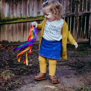 May include: A young child wearing a white shirt with the text "We rise by lifting others" and a yellow cardigan, holds a rainbow streamer toy. The child is wearing blue denim shorts and yellow tights. The child is standing on a dirt path.