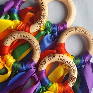 Op de afbeelding: Vier houten ringen met regenboog lint streamers. Elke ring heeft een gegraveerde naam: Coralie, Mouse loves Pig, Madison en Noah. De Noah ring heeft een spinnenweb en een spin ontwerp.