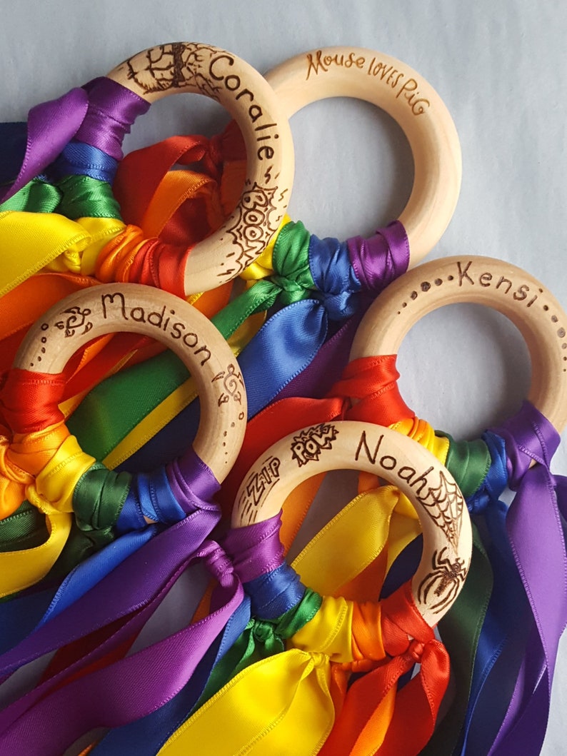 Op de afbeelding: Vier houten ringen met regenboog lint streamers. Elke ring heeft een gegraveerde naam: Coralie, Mouse loves Pig, Madison en Noah. De Noah ring heeft een spinnenweb en een spin ontwerp.