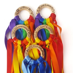 Op de afbeelding: Vijf houten ringen met regenboogkleurige linten eraan bevestigd. Elke ring heeft een andere naam erop geschreven: William, Jon Lucas, Emmerson, Liam en Elliot.