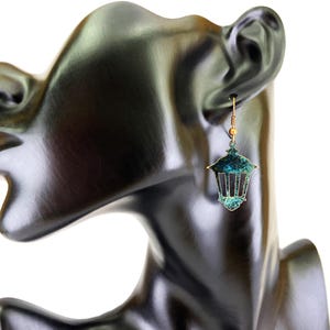 May include: A pair of dangle earrings featuring a teal blue and gold lantern design. The earrings are hanging from a mannequin's ear.