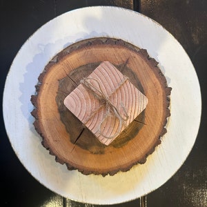 Puede incluir: Un pequeño cuadrado de madera envuelto en cuerda se encuentra sobre una rodaja de madera, que está sobre un plato blanco. La rodaja de madera tiene un patrón de veta de madera natural.