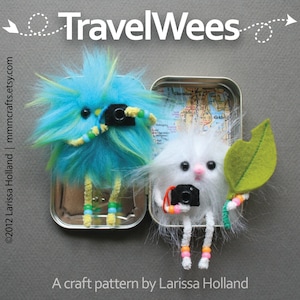 May include: Two whimsical, furry creatures, one blue and one white, each holding a tiny camera. They are inside a small metal tin with a map. The text "Travel Wees" and "A craft pattern by Larissa Holland" are also visible.