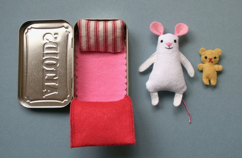 May include: A white mouse with pink ears and a red nose is lying in a small bed made from a tin Altoids container. The bed has a pink and red fabric lining and a white and red striped pillow. A small yellow teddy bear is standing next to the bed.