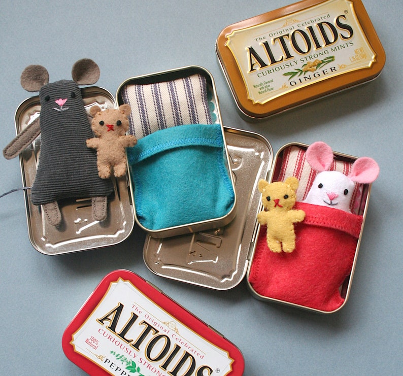 May include: Three small, handmade stuffed animals, a mouse, a bear, and a rabbit, are placed inside empty Altoids mint tins. Each tin has a different colored fabric lining and a different flavor of Altoids mint tin. The tins are labeled "The Original Celebrated Altoids Curiously Strong Mints" with the flavors "Ginger", "Peppermint", and "Wintergreen".
