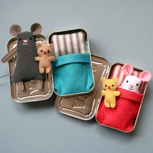 May include: Three small tin containers with lids, each containing a stuffed animal and a fabric bed. The first tin has a gray mouse with a red nose and a brown teddy bear. The second tin has a blue fabric bed with white stripes. The third tin has a red fabric bed with white stripes and a white mouse with a pink nose and a yellow teddy bear.