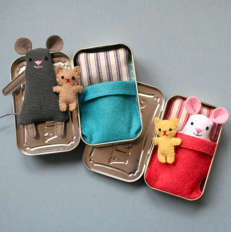 May include: Three small tin containers with lids, each containing a stuffed animal and a fabric bed. The first tin has a gray mouse with a red nose and a brown teddy bear. The second tin has a blue fabric bed with white stripes. The third tin has a red fabric bed with white stripes and a white mouse with a pink nose and a yellow teddy bear.
