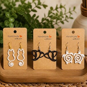 Puede incluir: Tres pares de pendientes hechos a mano en tarjetas marrones. Los pendientes presentan diseños de gato, pájaro y tortuga en blanco y negro. Cada tarjeta tiene el texto "Handmade with love".