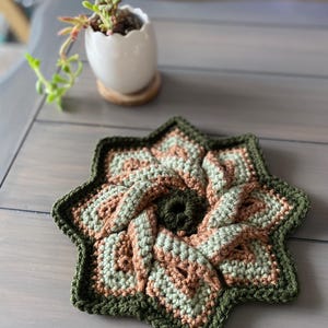 May include: A crocheted star-shaped coaster in shades of green and brown. The coaster is made with a spiral pattern and has a textured surface.