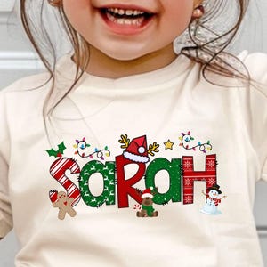 May include: A toddler's cream-colored Christmas t-shirt features the name 'Sarah' in colorful, festive letters. Each letter is decorated with various Christmas elements like candy canes, holly, snowflakes, a Santa hat, a gingerbread man, and a snowman. The shirt is a comfortable, short-sleeved style, perfect for the holiday season.