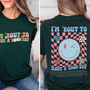 Puede incluir: Camiseta verde oscuro con la frase "I'M 'BOUT TO HAVE A GOOD DAY" en letras coloridas de estilo retro. La parte trasera de la camiseta presenta una carita sonriente gui&ntilde;ando un ojo, flores y la misma frase, sobre un fondo a cuadros rosa y blanco.