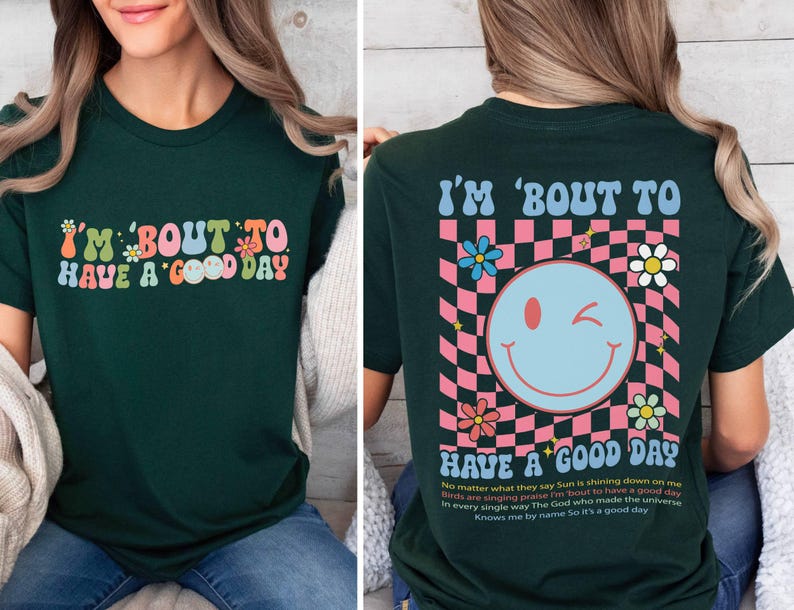 Puede incluir: Camiseta verde oscuro con la frase "I'M 'BOUT TO HAVE A GOOD DAY" en letras coloridas de estilo retro. La parte trasera de la camiseta presenta una carita sonriente gui&ntilde;ando un ojo, flores y la misma frase, sobre un fondo a cuadros rosa y blanco.