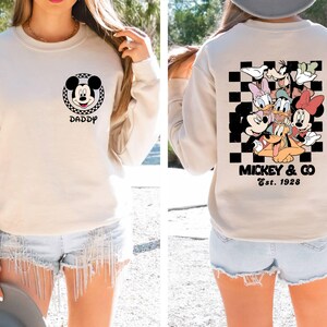 Puede incluir: Una sudadera beige con un patr&oacute;n de cuadros blanco y negro y un gr&aacute;fico de Mickey Mouse en la parte delantera. La parte posterior de la sudadera tiene un patr&oacute;n de cuadros blanco y negro con Mickey Mouse y sus amigos. El texto "Mickey & Co. Est. 1928" est&aacute; impreso en la parte posterior de la sudadera.