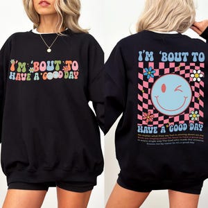 May include: Black sweatshirt with the text "I'M 'BOUT TO HAVE A GOOD DAY" in colorful, retro font. The back features a smiley face graphic on a pink and white checkered background, with additional text and floral accents.