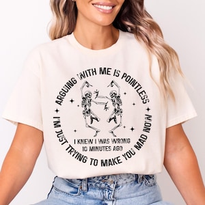May include: Cream-colored t-shirt with a black graphic of two dancing skeletons inside a circular design. The text around the skeletons reads "ARGUING WITH ME IS POINTLESS, I'M JUST TRYING TO MAKE YOU MAD NOW, I KNEW I WAS WRONG 10 MINUTES AGO."