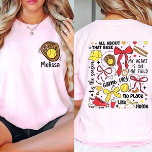 May include: Pink t-shirt with a softball and glove graphic on the front and a design with the text "All About That Base", "Tis the Season", "Game Day", "My Heart is on That Field", "No Place Like Home" on the back.