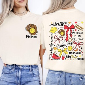 May include: A cream-colored t-shirt with a softball-themed design. The front of the shirt has a cartoon image of a glove catching a softball. The back of the shirt has a variety of softball-related images, including a baseball cap, a glove, a trophy, a drink, a bow, and a baseball. The text on the back of the shirt reads "All About That Base", "Tis The Season", "Game Day", "My Heart Is On That Field", "No Place Like Home".
