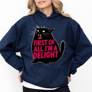 Op de afbeelding: Marineblauwe hoodie met een zwarte kattenafbeelding en de roze tekst "FIRST OF ALL, I'M A DELIGHT". De kat heeft een open mond. De sweatshirt heeft een voorzak en een capuchon met trekkoord. Een casual, comfortabel kledingstuk.
