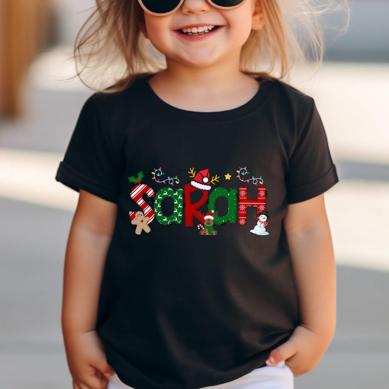 Op de afbeelding: Zwart T-shirt met een kerstthema ontwerp met de naam "Sarah" in kleurrijke letters versierd met feestelijke elementen zoals snoepstokken, lichtjes en een sneeuwpop.