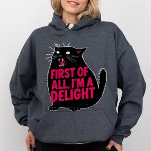 Op de afbeelding: Donkergrijze hoodie met een zwarte kattenillustratie en de roze tekst "FIRST OF ALL, I'M A DELIGHT". De kat heeft witte snorharen en rode tanden. De hoodie heeft een voorzak en een capuchon.