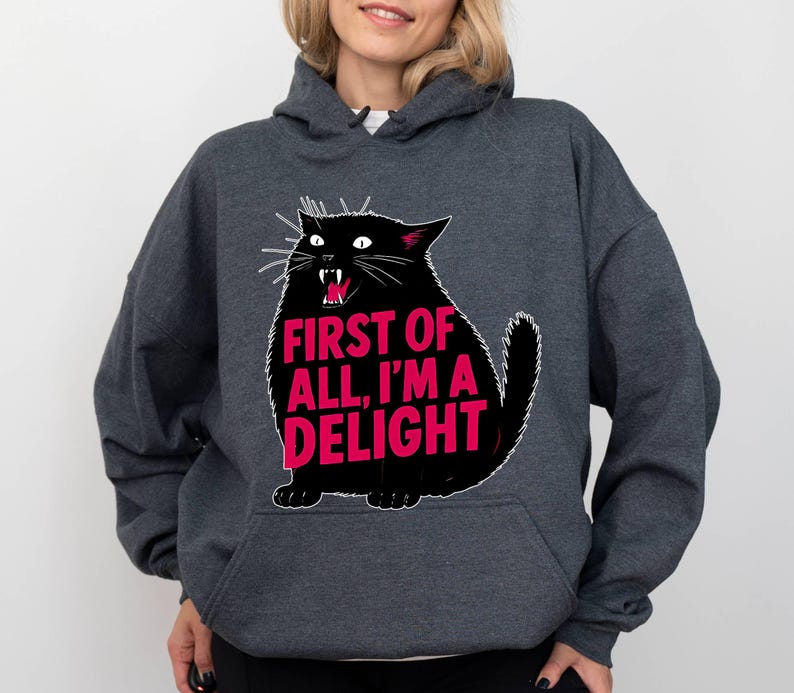 Op de afbeelding: Donkergrijze hoodie met een zwarte kattenillustratie en de roze tekst "FIRST OF ALL, I'M A DELIGHT". De kat heeft witte snorharen en rode tanden. De hoodie heeft een voorzak en een capuchon.
