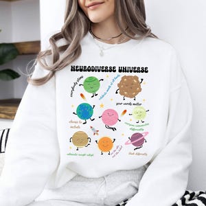K&ouml;nnte beinhalten: Wei&szlig;er Pullover mit einem grafischen Design aus Planeten und Sternen. Der Text "Neurodiverse Universe" ist in einer Retro-Schrift gedruckt. Die Planeten haben Gesichter und sind von S&auml;tzen wie "Jeder strahlt", "Feiert die K&ouml;pfe aller Arten", "Deine Worte z&auml;hlen", "Jeder kommuniziert anders", "Seht die F&auml;higkeit, nicht das Etikett", "Denkt anders", "W&auml;hlt das Einschlie&szlig;en", "Setzt euch ein, akzeptiert, passt euch an" umgeben.