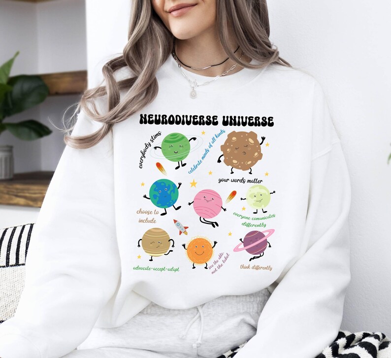 K&ouml;nnte beinhalten: Wei&szlig;er Pullover mit einem grafischen Design aus Planeten und Sternen. Der Text "Neurodiverse Universe" ist in einer Retro-Schrift gedruckt. Die Planeten haben Gesichter und sind von S&auml;tzen wie "Jeder strahlt", "Feiert die K&ouml;pfe aller Arten", "Deine Worte z&auml;hlen", "Jeder kommuniziert anders", "Seht die F&auml;higkeit, nicht das Etikett", "Denkt anders", "W&auml;hlt das Einschlie&szlig;en", "Setzt euch ein, akzeptiert, passt euch an" umgeben.