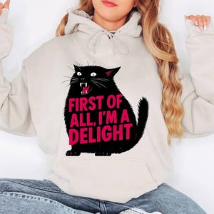 Op de afbeelding: Een beige hoodie met een zwarte kattenafbeelding en de roze tekst "FIRST OF ALL, I'M A DELIGHT". De kat heeft een open mond en scherpe tanden. De hoodie heeft een voorzak en een capuchon met trekkoord.