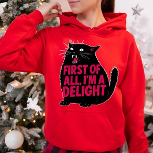 Op de afbeelding: Een rode hoodie met een zwarte kattenafbeelding en de roze tekst "FIRST OF ALL, I'M A DELIGHT". De kat heeft een open mond en witte snorharen. De hoodie heeft een voorzak.