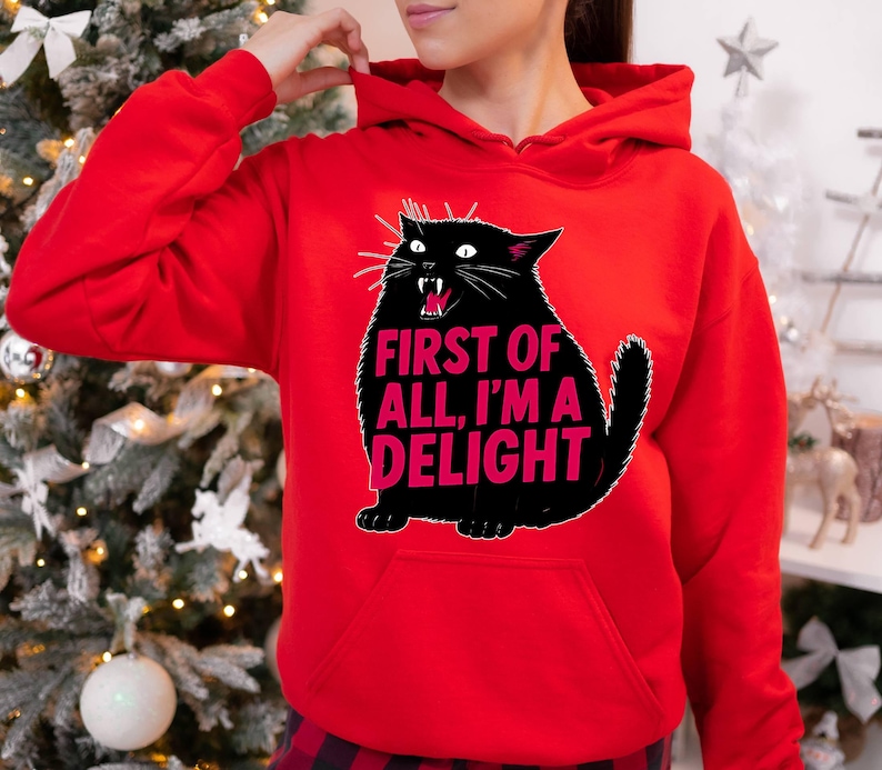 Op de afbeelding: Een rode hoodie met een zwarte kattenafbeelding en de roze tekst "FIRST OF ALL, I'M A DELIGHT". De kat heeft een open mond en witte snorharen. De hoodie heeft een voorzak.