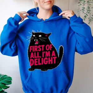 Op de afbeelding: Koningsblauwe hoodie met een zwarte kattenafbeelding. De kat heeft een open mond en de tekst "FIRST OF ALL, I'M A DELIGHT" in roze. De hoodie heeft een capuchon met trekkoord.